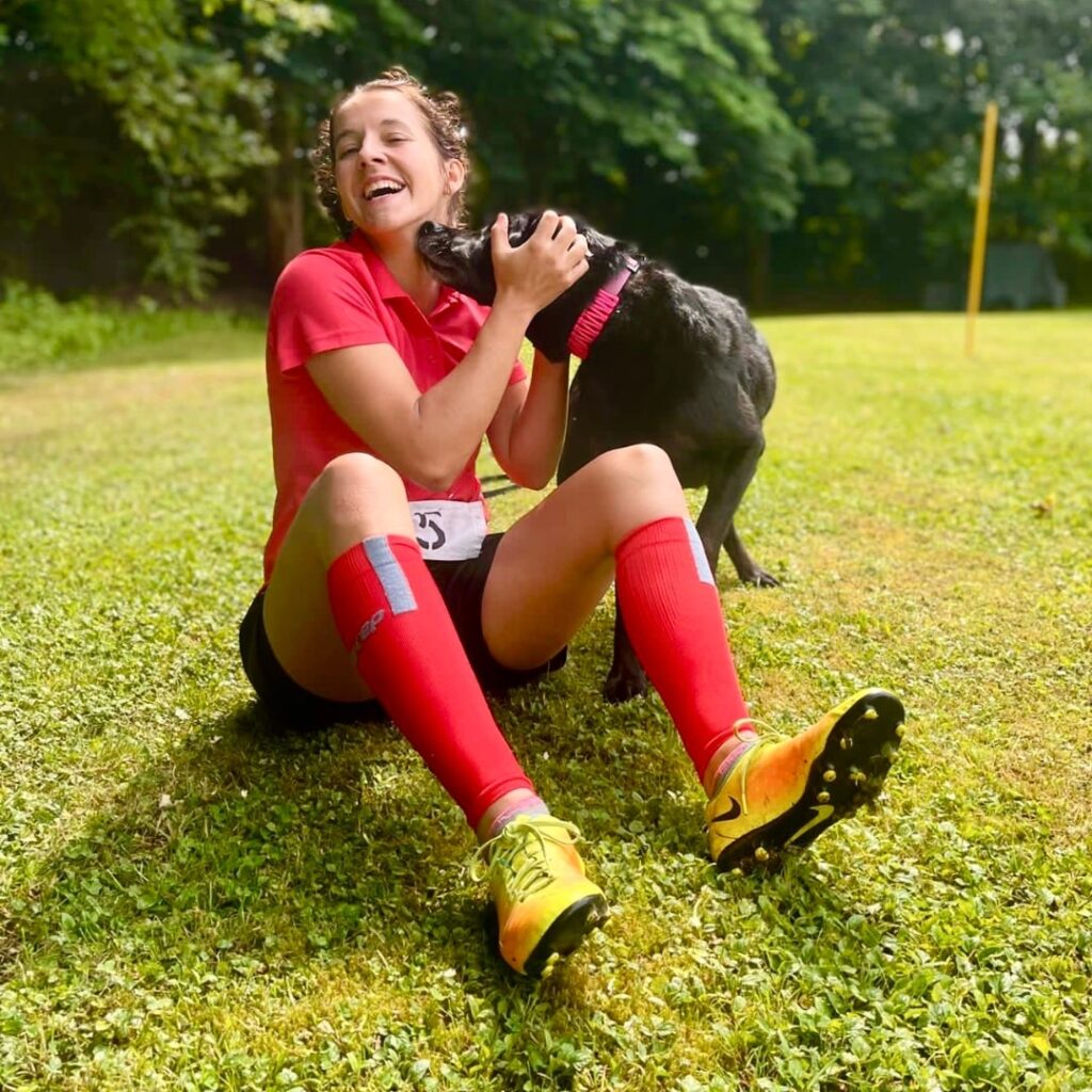 Julia Pöschel sitzt lachend auf dem Boden und streichelt Labrador Balu während dem Turnierhundesport Training