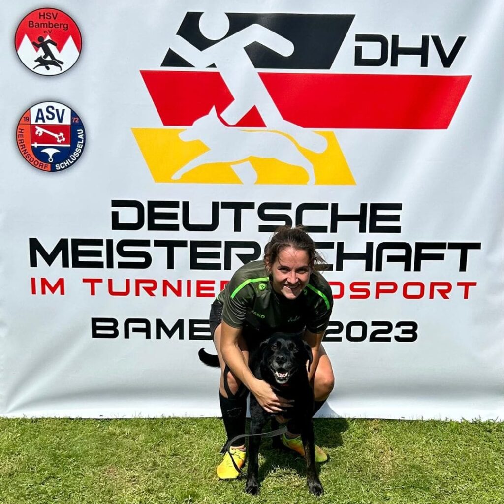 THSlerin Julia Pöschel steht vor dem Banner zur Deutschen Meisterschaft im Turnierhundsport gemeinsam mit ihrem schwarzen Labrador Balu lächelt sie in die Hundesportlerin in die Kamera.