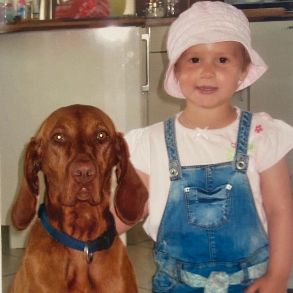 Lilien Hauser umarmt als kleines Kind den damaligen Familien- und Sporthund, einen Magyar Vizsla, und schaut lachend in die Kamera.